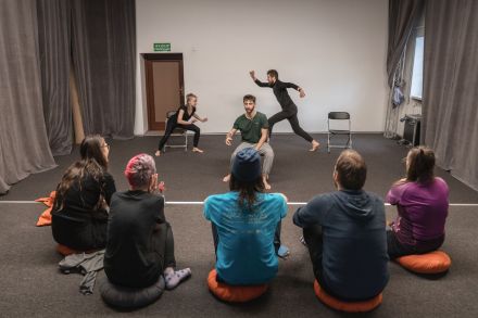 Po scenie biega mężczyzna, a kobieta i mężczyzna siedzą na krześle. Przygląda im się pięć osób.