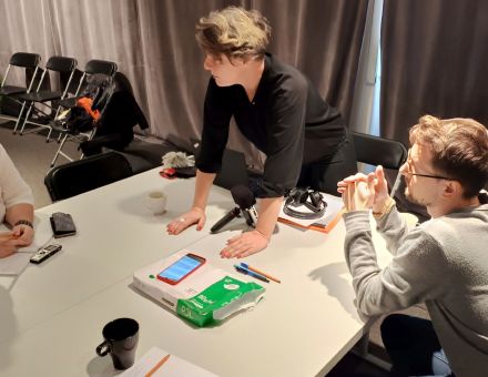 Uczestnicy warsztatów siedzą przy stole, na którym leżą telefony i dyktafony. Prowadząca stoi oparta o blat.