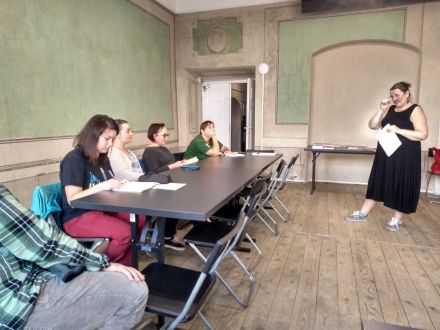 Uczestniczki notują coś przy stole, a prowadząca do nich mówi.