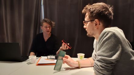 Kobieta i mężczyzna siedzą przy stole. Trzymają w ręce telefony i mówią do mikrofonów.