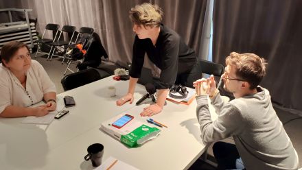 Uczestnicy warsztatów siedzą przy stole, na którym leżą telefony i dyktafony. Prowadząca stoi oparta o blat.