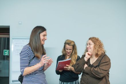 Grupa trzech kobiet stoi na środku i prezentuje coś przed grupą. Jedna z nich mówi, druga się zastanawia, a trzecia robi notatki.