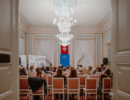 Zdjęcie sali, w której odbywa się wieczorek autorski. Sala pełna krzeseł, bardzo duży żyrandol, na ścianie godło Polski oraz logo Warsztatów Kultury. W głębi stół dla gości i prowadzących.