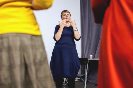 Na zdjęciu widzimy prowadzącą, która przybliża dłonie do twarzy i tłumaczy coś uczestniczkom.