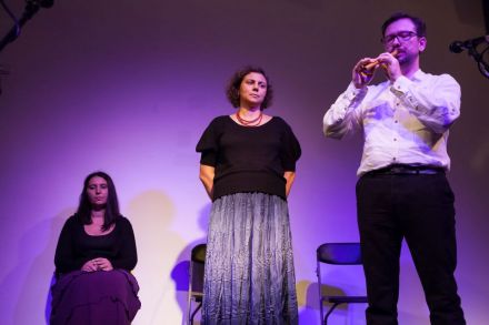 Na zdjęciu stoi mężczyzna grający na flecie, obok niego stoi jedna ze śpiewaczek. Druga siedzi pod ścianą.