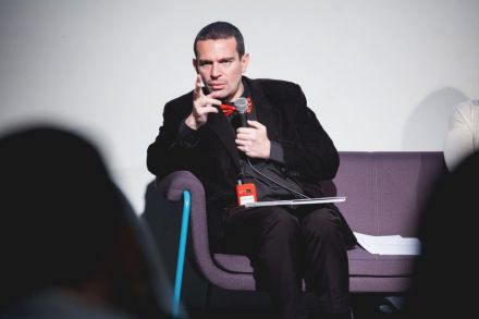 Na białym tle sceny na fioletowej kanapie siedzi mężczyzna, który znajduje się w centrum zdjęcia. Jest elegancko ubrany, wskazując palcem zwraca się do zebranej publiczności.