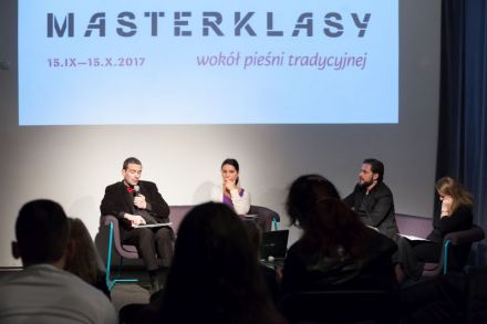 Czwórka elegancko ubranych osób siedzi w fotelach i kanapach na scenie. Za nimi wyświetlany jest napis \"MASTERKLASY wokół pieśni tradycyjnej\". Zdjęcie zrobione zza pleców publiczności zgromadzonej naprzeciwko biorących udział w dyskusji panelistów.