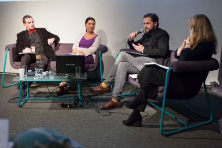 Zbliżenie na cztery osoby siedzące na kanapach rozłożonych na scenie. Przed nimi stoi stolik ze szklankami wody i otwarty laptop. Dwóch mężczyzn, dwie kobiety. Jeden z mężczyzn ma w ręku mikrofon i zwraca się w stronę publiczności.