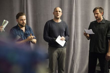 Na zdjęciu widzimy trzech mężczyzn w tym prowadzącego oraz dwóch uczestników, którzy trenują techniki wokalne. Jeden zerka na tekst, drugi patrzy przed siebie.