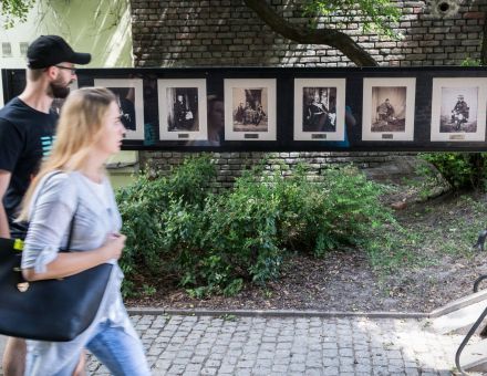 Dwie osoby, mężczyzna i kobieta, ida wzdłuż wystawy. Za nimi widać rząd czarno-białych zdjęć w białych ramkach,