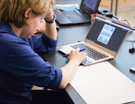 Kobieta siedzi przy biurku, jedną ręką podpiera głowę, drugą pisze na klawiaturze laptopa. Z prawej strony komputera biała kartka papieru.