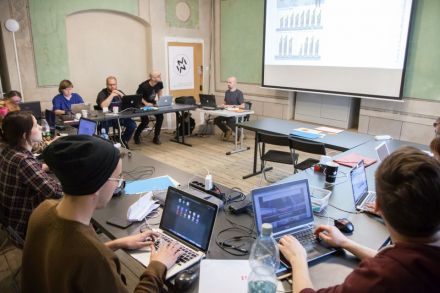 Zdjęcie przedstawia widok na całą salę MediaLabową Warsztatów Kultury. Dookoła  na kształt podkowy rozłożone są biurka przy których zgromadzona jest grupa uczestników warsztatów.  Wszyscy mają otworzone przed sobą laptopy. Na przeciwko znajduje się duży ekran rzutnika.