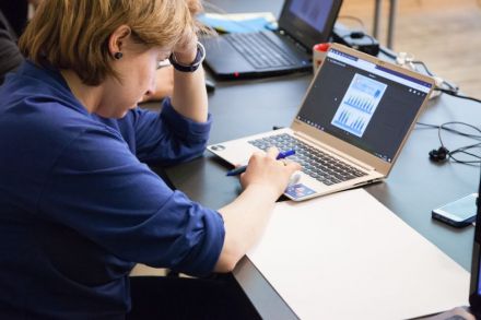 Kobieta siedzi przy biurku, jedną ręką podpiera głowę, drugą pisze na klawiaturze laptopa. Z prawej strony komputera biała kartka papieru.