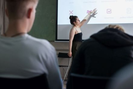Zdjęcie zrobione zza pleców dwóch warsztatowiczów siedzących na krzesłach. Pomiędzy nimi widzimy ekran rzutnika i prowadzącą warsztaty młodą kobietę, wskazującą na rzut ekranu.