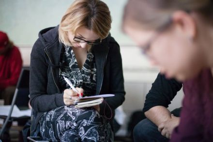 Zbliżenie na jedną z uczestniczek warsztatu. Jest to młoda kobieta,  siedzi na krześle i notuje coś w notesie leżącym na jej kolanie.
