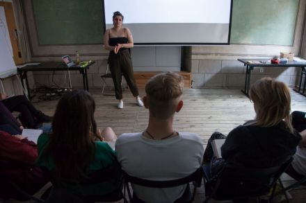 Zdjęcie zza pleców osób siedzących w pierwszym rzędzie na szkoleniu.  Widoczna prowadząca i wiszący za jej plecami ekran.