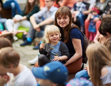 Na środku zdjęcia znajduje się młoda kobieta pochylona nad małym dzieckiem. Oboje się uśmiechają.  Znajdują się oni pośród szerokiego grona publiczności koncertu \"Jerz Igor\".