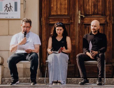 Na zdjęciu widać trzy osoby siedzące przed ratuszem. Jedną z osób jest Bogusław Wróblewski z czasopisma Akcent, trzyma on mikrofon i odczytuje treść z kartki.