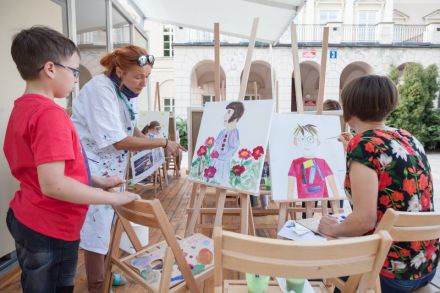 Na zdjęciu widzimy prowadzącą przyglądającą się obrazowi chłopca. Namalował on siedzącą obok kobietę pośród kwiatów.