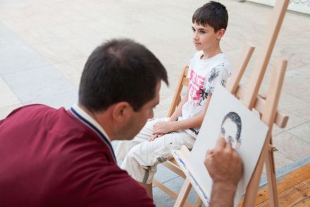 Na zdjęciu widać mężczyznę malującego ze skupieniem chłopca siedzącego obok niego na krześle. Pozuje, sam nie maluje obrazu.
