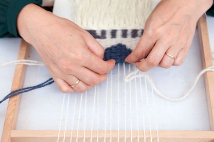 Kompozycja przedstawia kobiece dłonie dociskające kilim na podręcznym krośnie. Kilim jest w fazie początkowej tworzenia. Nadgarstki lekko przykrywa rękaw szmaragdowej koszuli. Wszystko na białym tle stołu.