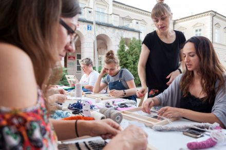 Grupa młodych kobiet zebrana wokół stołu na placu Litewskim zabiera się do pracy. Na stole panuje przyjemny chaos. Kobiety wydają się być zainteresowane, są kolorowo ubrane. Dzień jest jasny i ciepły.