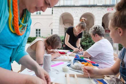 Kobiety zebrane wokół stołu oddają się pracy nad wyplataniem kilimów. W tle krzewy i zabudowa architektoniczna jednego z budynków UMCS.  Jest jasno, pogoda jest letnia.