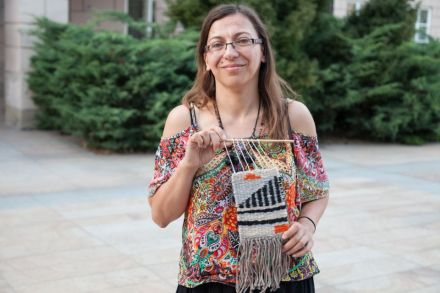 Uśmiechnięta uczestniczka warsztatów przedstawia zrobiony przez siebie kilim. Kobieta jest młoda, ma na sobie kolorową koszulkę. Kilim trzyma na wysokości klatki piersiowej.