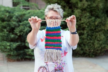 Kobieta stojąca na zielonym tle krzewów trzyma na wysokości twarzy wypleciony przez siebie kilim w różnokolorowe paski.