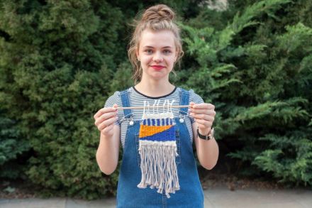 Młoda uśmiechnięta kobieta w ogrodniczkach, stoi na tle zielonych krzewów i przedstawia przed obiektywem zrobiony przez siebie kilim.