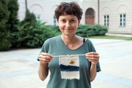 Młoda kobieta trzyma w ręku kilim wykonany podczas warsztatów. Kobieta jest uśmiechnięta, stoi na tle wydziału politologii UMCS na placu Litewskim.