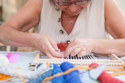 Zbliżenie na twarz, i ręce kobiety zaczynającej wyplatać kilim na małym krośnie leżącym na stole. Przed nią leżą kolorowe motki muliny.