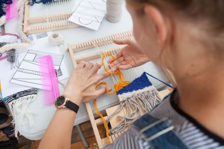 Zdjęcie przedstawia widok zza pleców dziewczyny wyplatającej kilim. Kilim który powstaje jest w kolorach żółto-niebieskich. Obok krosna leży różowy grzebień. Krosno leży na białym stole, który jest tłem zdjęcia.