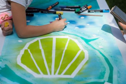 Na zdjęciu widzimy ręce dziewczynki, która kończy malować obraz. Widocznym motywem obrazu jest wielka żółto-zielona muszelka.