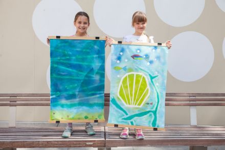Na zdjęciu dwie dziewczynki stojące obok siebie i trzymające swoje skończone prace. Pierwszy obraz przedstawia plażę i bezkres wody, a drugi wielką muszelkę i rybki na dnie morza.