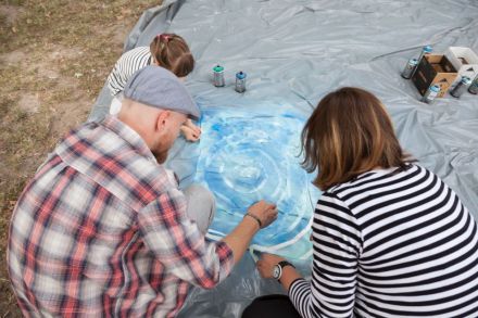 Na zdjęciu mężczyzna razem z kobietą pomagają przy tworzeniu dzieła jednej z dziewczynek. Układają spiralnie taśmę na pomalowanym na niebiesko płótnie.