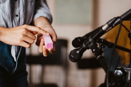 Z prawej strony zdjęcia dwa mikrofony na statywach, z lewej dłonie trzymające w ręku różowy podłużny kubik. Jedna ręka trzyma przedmiot, druga ręka przesuwa po nim palcem.