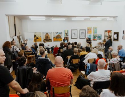 Zdjęcie zrobione z tyłu sali, zza pleców publiczności. Widać wszystkich zgromadzonych ludzi oraz prowadzącą i gościa spotkania. Prowadząca, pani Jadwiga Mizińska, stoi i przemawia do publiczności.