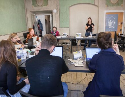 Na zdjęciu widać uczestników siedzących przed laptopami, naprzeciwko nich stoi prowadząca.