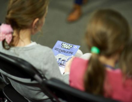 Zdjęcie zrobione od tyłu, zbliżenie na dwie dziewczynki, jedna z nich trzyma na kolanach komiks, związany z tematyką spotkania.