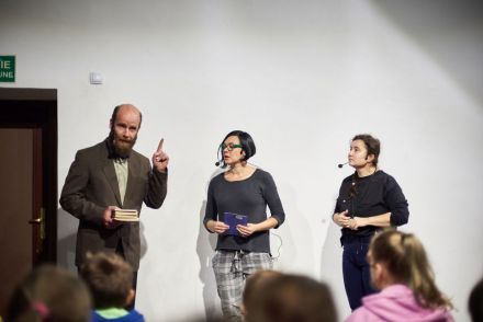 Zdjęcie ukazuje trzy osoby. Do duetu aktorskiego dołączył kolejny pracownik Warsztatów Kultury. Mężczyzna trzyma w ręce książki i mówi do dzieci.