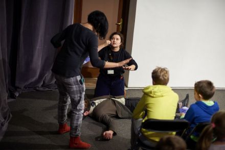 Na zdjęciu widać leżącego mężczyznę oraz dwie koordynatorki obok niego, próbujące zachować spokój. Widzimy dwóch chłopców z publiczności siedzących najbliżej rozgrywanej akcji.