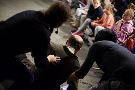 Na zdjęciu kobiety próbują pomóc mężczyźnie wstać. Dzieci z zainteresowaniem przyglądają się sytuacji.