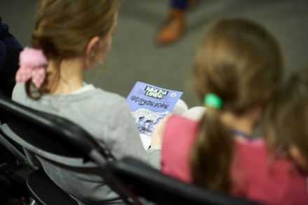 Zdjęcie zrobione od tyłu, zbliżenie na dwie dziewczynki, jedna z nich trzyma na kolanach komiks, związany z tematyką spotkania.