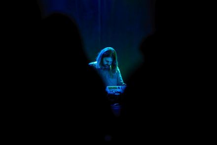 Zdjęcie zrobione zza pleców osób z publiczności. Pomiędzy ciemnymi, niewyraźnymi zarysami publiczności widać artystę.