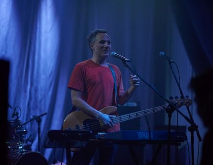 Na środku zdjęcia w zacienionej sali koncertowej stoi artysta z gitarą basową.