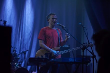 Na środku zdjęcia w zacienionej sali koncertowej stoi artysta z gitarą basową.