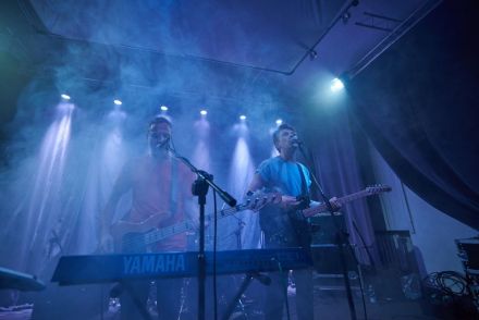 Zdjęcie wykonane z żabiej perspektywy. Dwóch muzyków stojących w zadymionej sali koncertowej, w ich tle reflektory rzucające światło na scenę. Przed nimi stoją klawisze.