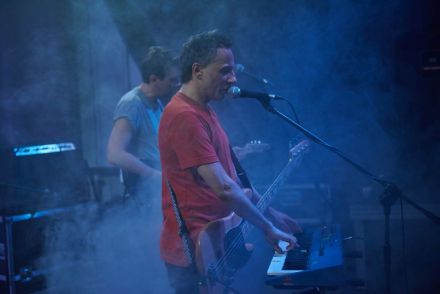 Zdjęcie przedstawiające muzyka grającego na klawiszach z przewieszoną przez szyję gitarą basową. Muzyk równocześnie śpiewa do mikrofonu. W tle drugi członek zespołu Ukryte Zalety Systemu - gitarzysta.