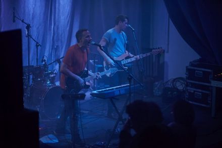 Dwóch muzyków na scenie, z gitarami w przewieszonymi przez szyje, przy ustach mają mikrofony. Przed jednym z nich stoją klawisze. Widok z boku.